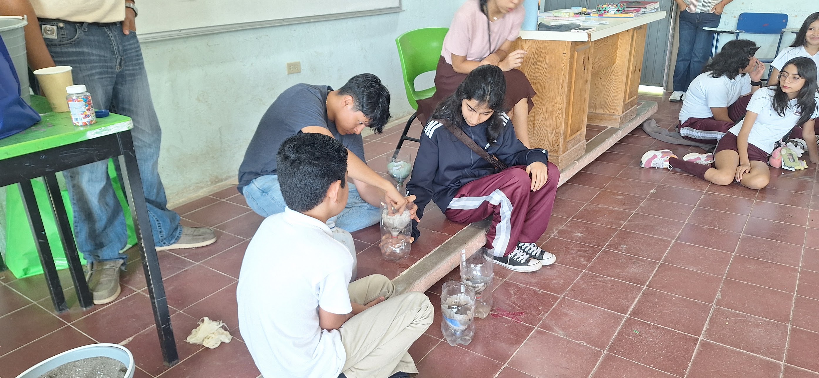 Children seated on tiled classroom floor gathered around plastic bottles and supplies