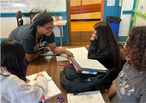 Mentor teacher smiling and leaning over to provide instruction to a student in a classroom.